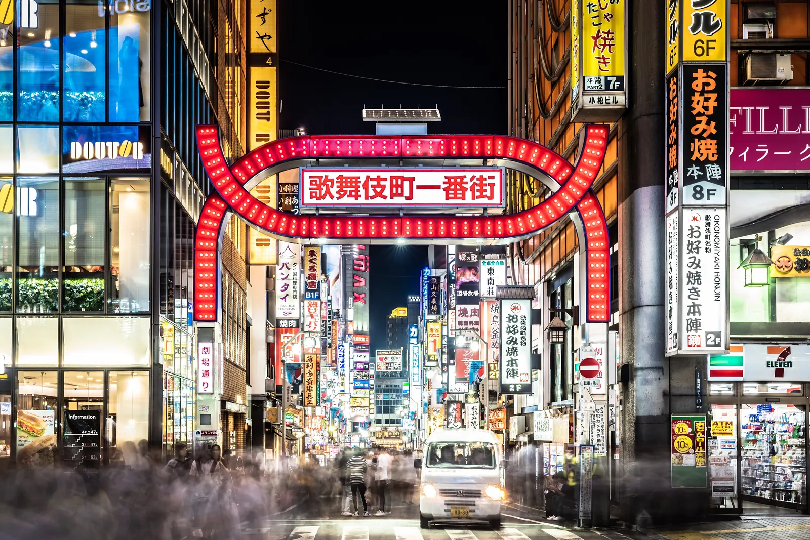 The red arched Kabukichō Ichibangai gate lit at night, with neon signs lining the street beyond and a white van at the crossing