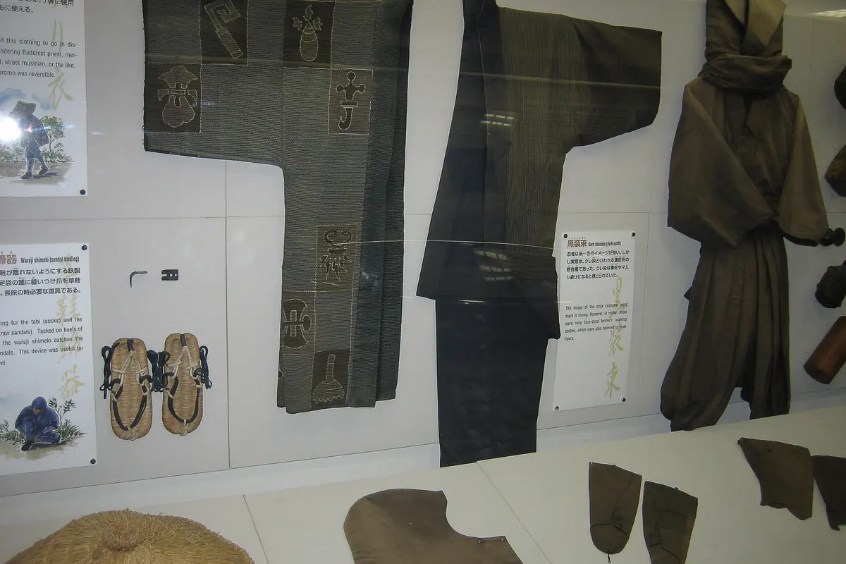 A museum case holding dark grey and brown work clothes, straw sandals and a hooded outfit, with printed labels beside each item