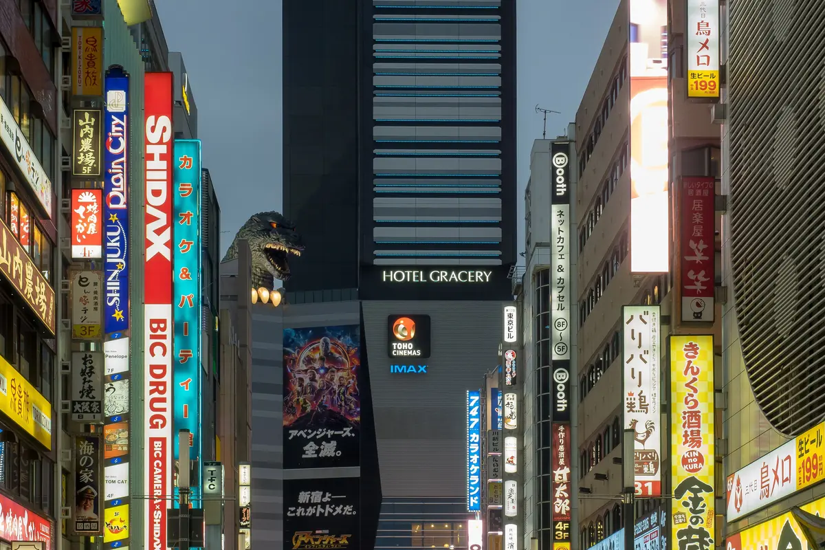 A narrow Shinjuku street of vertical signs at dusk with a Godzilla head looking over the rooftops beside a tall hotel tower