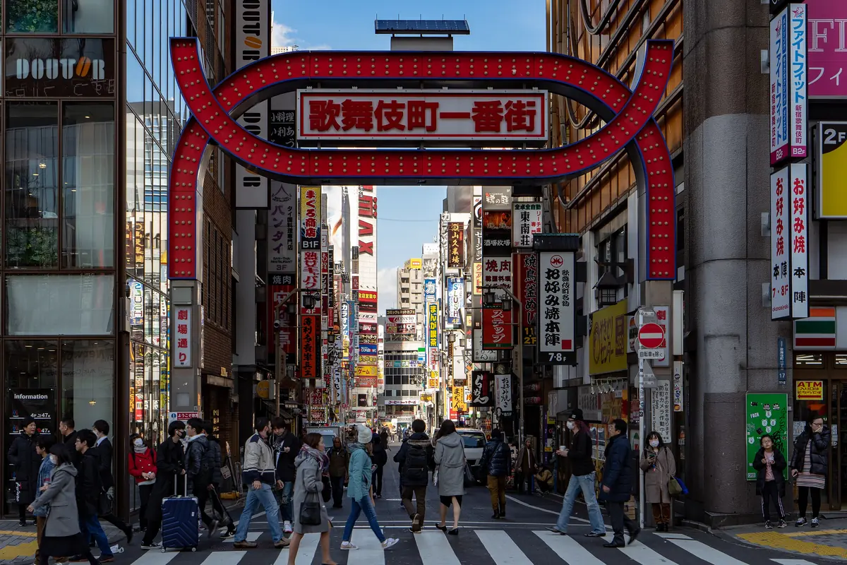 The red Kabukichō Ichibangai gate seen in daylight, with pedestrians crossing the road in front of tall signed buildings