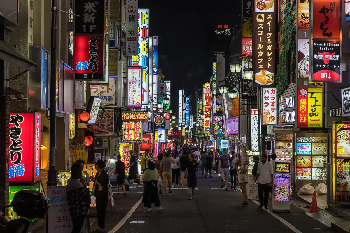 A narrow Shinjuku street at night lined with vertical illuminated signs in red, yellow and white, with crowds of pedestrians in the distance