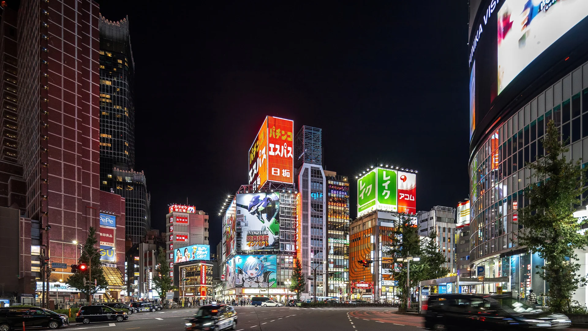 Illuminated towers with red, orange and green signs rise into a black night sky above a wide Shinjuku crossing with cars moving through it; an image of the district with no link to the performance