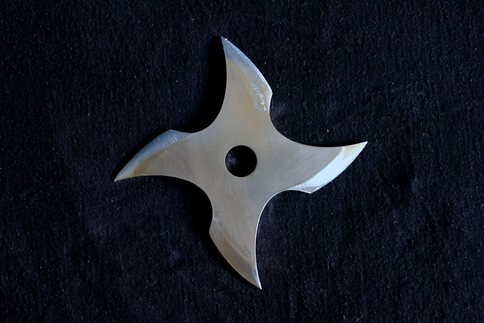 A four-pointed steel throwing star with a round hole at its centre on a dark cloth; a shuriken photographed as an object, not a prop from the show
